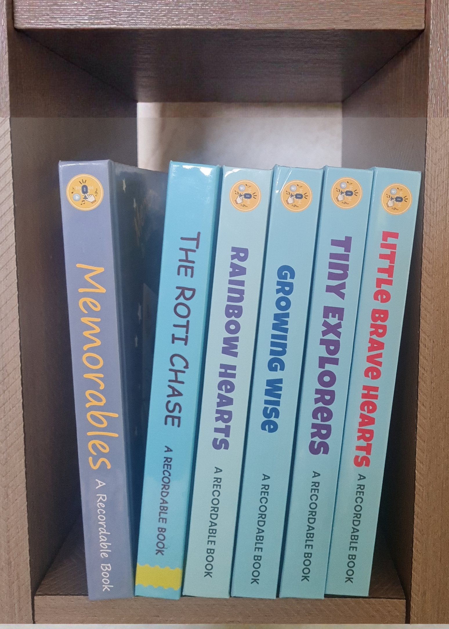 Set of books titled 'Memorables', 'The Rottl Chase', 'Rainbow Hearts', 'Growing Wise', and 'Tiny Brave Explorers' on a wooden shelf.